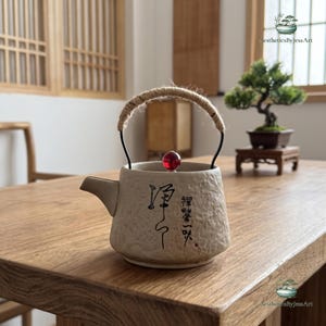 Può includere: Una teiera in ceramica beige con una superficie ruvida e irregolare e un beccuccio corto. Presenta un manico in metallo nero avvolto in corda color cuoio e un coperchio sormontato da una piccola perla rossa rotonda e traslucida. Calligrafia giapponese.