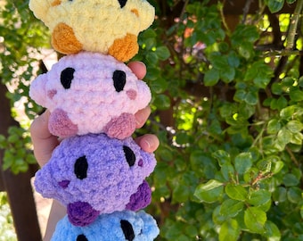 May include: A stack of four handmade crochet plush toys in yellow, pink, purple, and blue. Each plush has black eyes and contrasting colored feet. The toys are soft and fluffy, perfect for children or as decorative items.