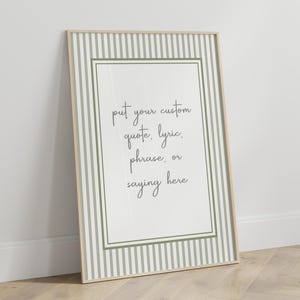 May include: A framed print with a light wood frame, leaning against a white wall. The print features a white background with a green and white striped border. The text on the print reads: "put your custom quote, lyric, phrase, or saying here."
