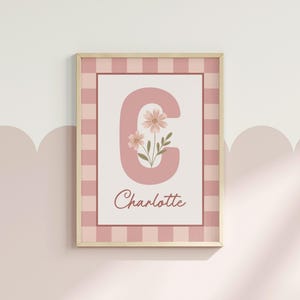 May include: A framed print with the letter 'C' in a soft pink colour, decorated with small flowers. The name 'Charlotte' is written below. The print is set within a pink and white checked border and a light wood frame.
