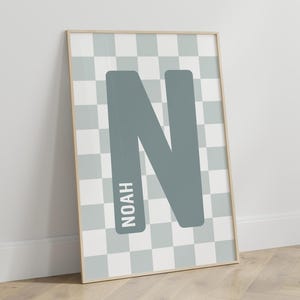 Puede incluir: Un estampado enmarcado con una gran letra "N" gris sobre un fondo a cuadros azul claro y blanco. El nombre "NOAH" está impreso verticalmente en el lado izquierdo de la letra. El marco es de color madera clara.