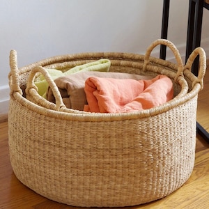 May include: A large, round, natural woven basket with two handles. The basket is filled with folded fabric in shades of green, brown, and orange.