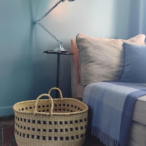 Puede incluir: Una cesta tejida con dos asas y un patrón geométrico negro. Una lámpara articulada plateada está montada en la pared, y un sofá con almohadas y una manta azul está en el fondo. La cesta tiene dos asas.