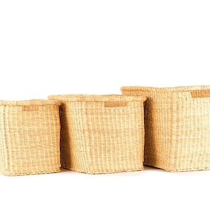 Pode incluir: Três cestos de arrumação tecidos em tamanhos variados. Os cestos são retangulares e de cor castanho-claro natural. Cada cesto tem uma pega retangular recortada na borda superior. Os cestos são feitos de um material tecido firmemente.