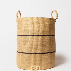 Puede incluir: Una cesta grande tejida de forma natural con dos rayas negras y dos asas. La cesta está hecha de materiales naturales y tiene un color natural y terroso. La cesta está etiquetada con "DSG".