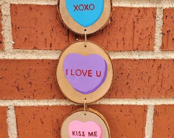 May include: Three wooden heart-shaped ornaments with painted messages. The top ornament is blue with the text "XOXO". The middle ornament is purple with the text "I LOVE U". The bottom ornament is pink with the text "KISS ME".