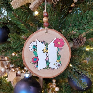 May include: A wooden Christmas ornament with a white painted "V" shape surrounded by colorful flowers. The ornament is hanging on a Christmas tree.