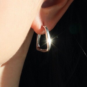May include: A close-up of a silver-coloured, geometric hoop earring. The earring has a rectangular shape with rounded edges. The earring is worn on the ear. The image is well-lit, highlighting the earring's metallic sheen.