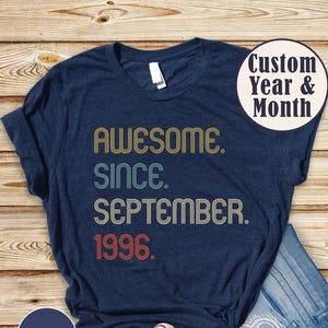 May include: A heather navy short-sleeve t-shirt featuring the text "AWESOME. SINCE. SEPTEMBER. 1996." in a retro, multi-colored font. The shirt is paired with blue denim jeans and white canvas sneakers, showcasing a casual, everyday ensemble.