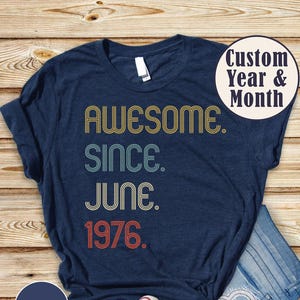 May include: Heather navy t-shirt with the text "AWESOME. SINCE. JUNE. 1976." in retro-style lettering. The shirt is paired with blue jeans and white sneakers. A circular graphic reads "Custom Year & Month".