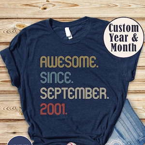 Op de afbeelding: Een heather navy t-shirt met de tekst AWESOME. SINCE. SEPTEMBER. 2001. in een retro lettertype met gele, lichtblauwe en rode kleuren. Een cirkelvormige afbeelding in de rechterbovenhoek toont de tekst Custom Year & Month.