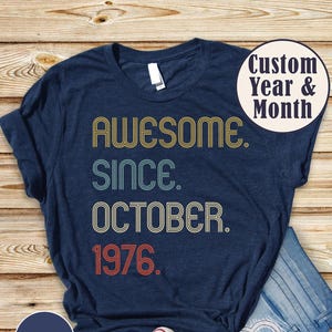 May include: A heather navy t-shirt featuring the text AWESOME SINCE OCTOBER 1976 in a retro, multi-line font with yellow, teal, and red outlines. A circular badge in the top right corner reads Custom Year & Month, and a badge in the bottom left reads HEATHER NAVY.