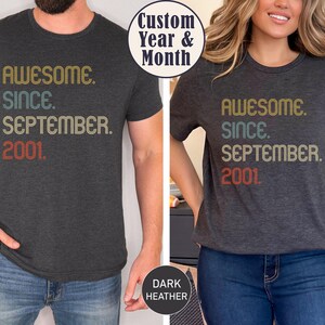 Op de afbeelding: Een donkergrijs gem&ecirc;leerd t-shirt met korte mouwen en de tekst "AWESOME. SINCE. SEPTEMBER. 2001." in gestapelde regels, gedrukt in een retro kleurenpalet van mosterdgeel, gedempt blauwgroen en vervaagd rood, met een ronde hals.