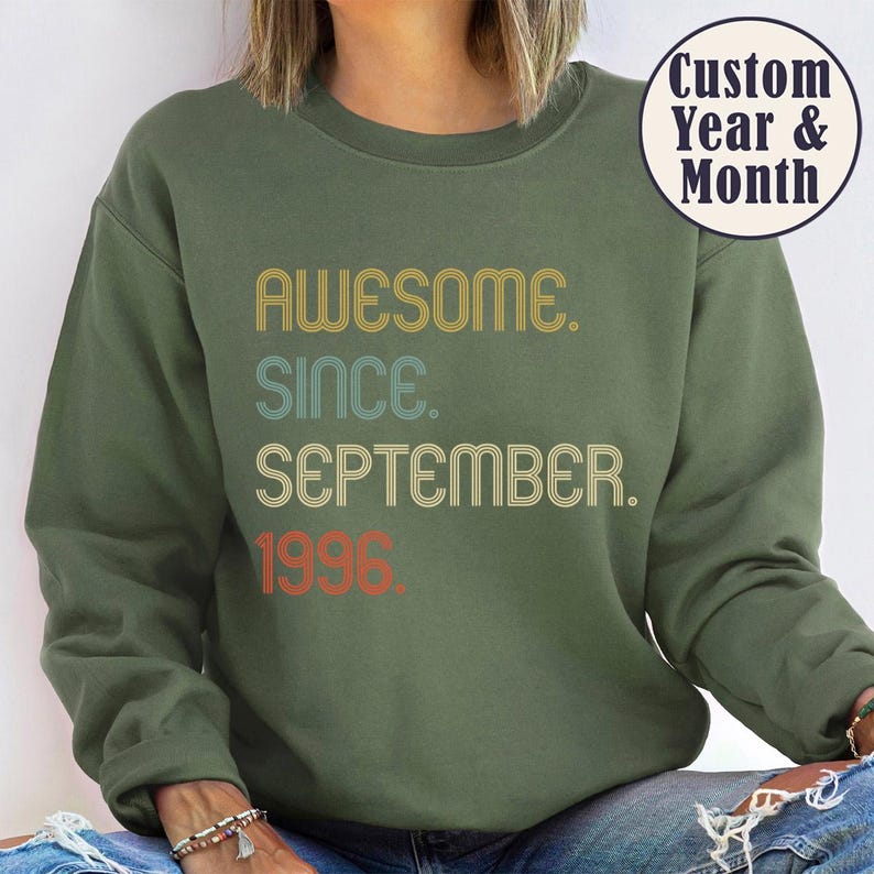 May include: An olive green crewneck sweatshirt featuring the text AWESOME. SINCE. SEPTEMBER. 1996. in a multi-colored, retro-style font. A circular badge in the upper right corner contains the text Custom Year & Month in dark blue lettering.