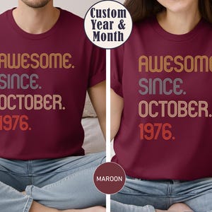 May include: A maroon short-sleeved t-shirt featuring the text "AWESOME. SINCE. OCTOBER. 1976." in a retro-style, multi-line font with orange, white, and grey striped outlines. The design is centered on the chest of the shirt.