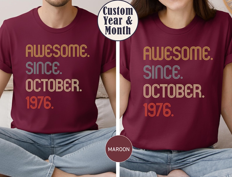 May include: A maroon short-sleeved t-shirt featuring the text "AWESOME. SINCE. OCTOBER. 1976." in a retro-style, multi-line font with orange, white, and grey striped outlines. The design is centered on the chest of the shirt.