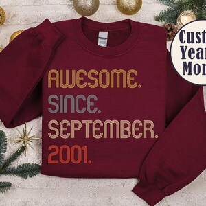 Op de afbeelding: Een bordeauxrode sweater met ronde hals en retro-stijl, meerregelige tekst in geel, grijs en oranje. De afbeelding toont AWESOME. SINCE. SEPTEMBER. 2001. Een cirkelvormige badge in de rechterbovenhoek toont de tekst Custom Year & Month.