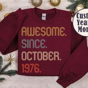 May include: A maroon long-sleeve crewneck sweatshirt featuring the text AWESOME. SINCE. OCTOBER. 1976. in a retro, multi-line font with orange, yellow, and grey colors. A circular badge in the top right corner displays the text Custom Year & Month.