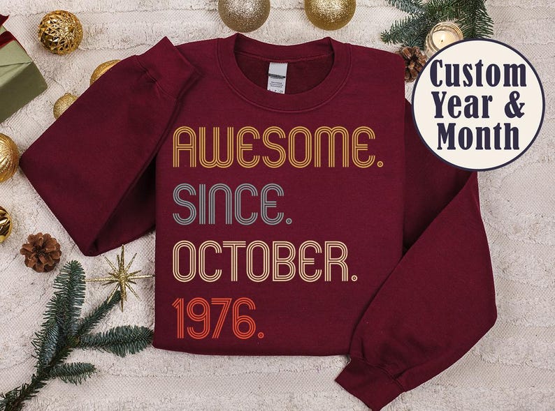 May include: A maroon long-sleeve crewneck sweatshirt featuring the text AWESOME. SINCE. OCTOBER. 1976. in a retro, multi-line font with orange, yellow, and grey colors. A circular badge in the top right corner displays the text Custom Year & Month.