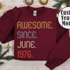 May include: Burgundy sweatshirt with the text "AWESOME. SINCE. JUNE. 1976." in retro-style lettering. The sweatshirt has long sleeves and a crew neck. A circular graphic reads "Custom Year & Month".