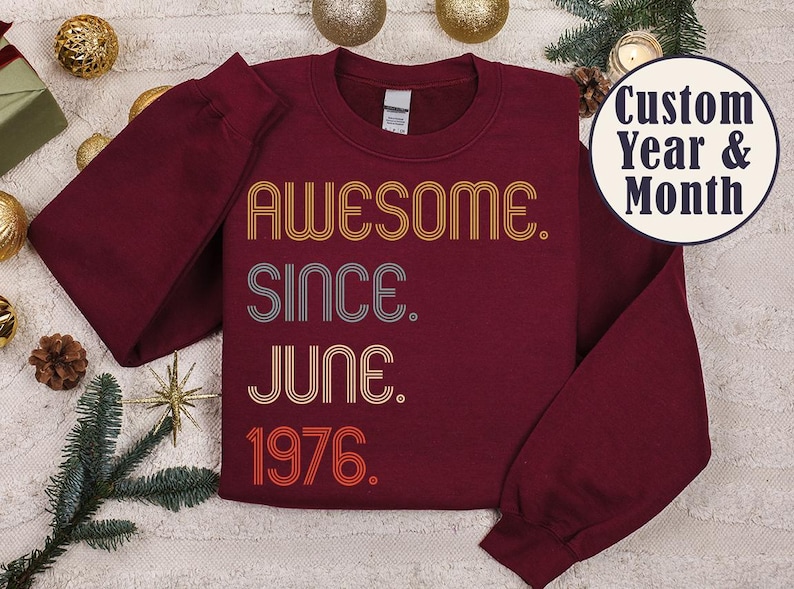 May include: Burgundy sweatshirt with the text "AWESOME. SINCE. JUNE. 1976." in retro-style lettering. The sweatshirt has long sleeves and a crew neck. A circular graphic reads "Custom Year & Month".