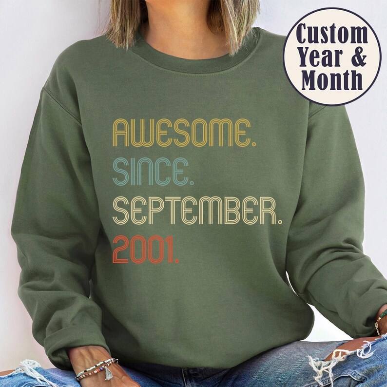 Op de afbeelding: Een olijfgroene sweater met ronde hals en de tekst AWESOME. SINCE. SEPTEMBER. 2001. in een retro lettertype met gele, blauwgroene en oranje letters. Een cirkelvormige badge in de rechterbovenhoek toont de tekst Custom Year & Month.
