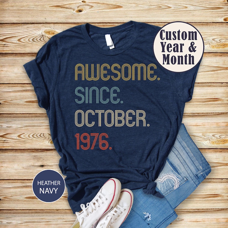May include: A heather navy t-shirt featuring the text AWESOME SINCE OCTOBER 1976 in a retro, multi-line font with yellow, teal, and red outlines. A circular badge in the top right corner reads Custom Year & Month, and a badge in the bottom left reads HEATHER NAVY.