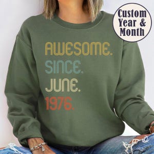 May include: Olive green sweatshirt with the text "AWESOME. SINCE. JUNE. 1976." in retro-style lettering. The sweatshirt has a crew neck and long sleeves. A circular graphic in the upper right corner reads "Custom Year & Month."