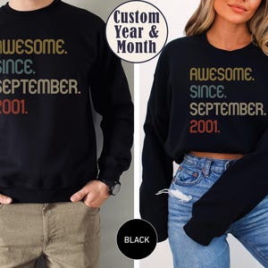 Op de afbeelding: Een zwart sweatshirt met lange mouwen met de tekst "AWESOME. SINCE. SEPTEMBER. 2001." in een retro-stijl lettertype met gouden, groenblauwe en rode letters. Het ontwerp wordt getoond in twee verschillende pasvormen, een standaard en een cropped lengte.