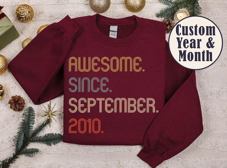 May include: A maroon long-sleeve crewneck sweatshirt featuring retro-style text in yellow, gray, and orange. The graphic reads AWESOME. SINCE. SEPTEMBER. 2010. A circular badge in the upper right corner displays the text Custom Year & Month.