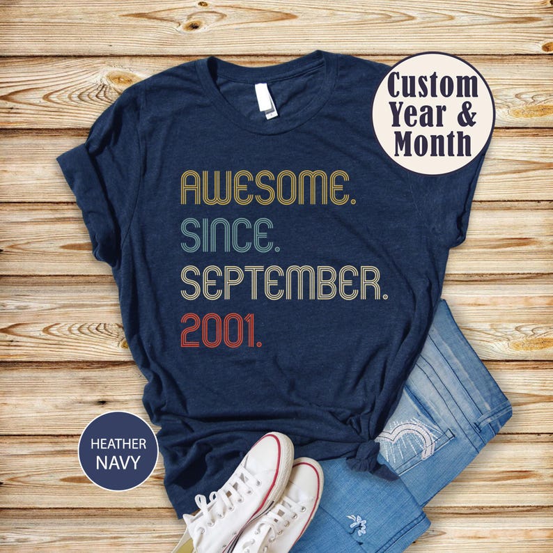 Op de afbeelding: Een heather navy t-shirt met de tekst AWESOME. SINCE. SEPTEMBER. 2001. in een retro lettertype met gele, lichtblauwe en rode kleuren. Een cirkelvormige afbeelding in de rechterbovenhoek toont de tekst Custom Year & Month.