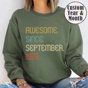 May include: An olive green crewneck sweatshirt featuring the text "AWESOME. SINCE. SEPTEMBER. 1976." in a retro, multi-colored font. A circular badge in the upper right corner displays the text "Custom Year & Month" in dark blue lettering.