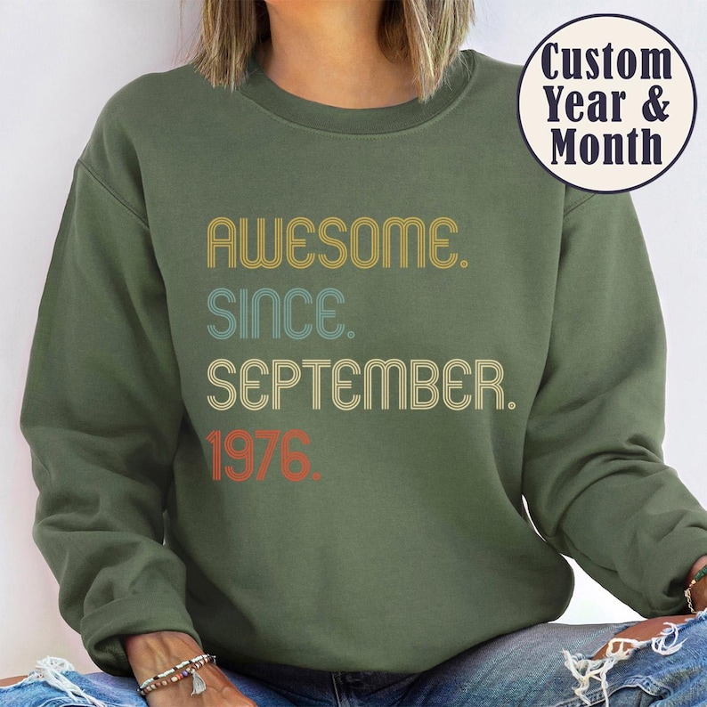 May include: An olive green crewneck sweatshirt featuring the text "AWESOME. SINCE. SEPTEMBER. 1976." in a retro, multi-colored font. A circular badge in the upper right corner displays the text "Custom Year & Month" in dark blue lettering.