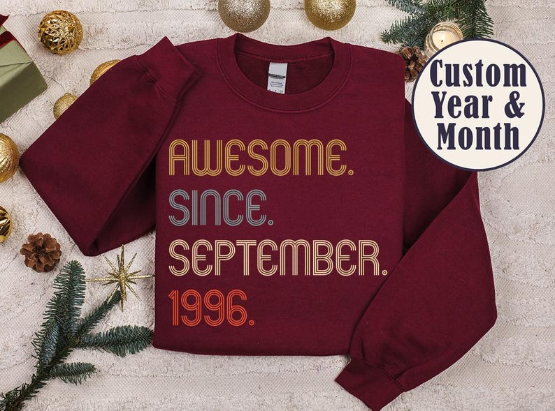 May include: A maroon long-sleeve crewneck sweatshirt featuring the text "AWESOME. SINCE. SEPTEMBER. 1996." in a retro, multi-line font with orange, grey, and red colors. The garment has a ribbed collar, cuffs, and hem, and a white interior neck label.