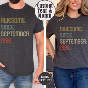 May include: A dark heather gray short-sleeve t-shirt featuring the text "AWESOME. SINCE. SEPTEMBER. 1996." printed in a retro-style font with gold, teal, and red colors. The shirt has a crew neckline and a standard fit, showcasing a vintage-inspired design.