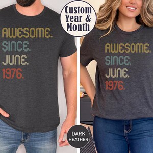 May include: Dark heather gray t-shirts with the text "AWESOME. SINCE. JUNE. 1976." in retro-style lettering. The shirts are worn by two people, one male and one female. The text is in yellow, teal, and red.