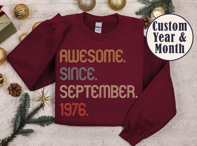 May include: A maroon crewneck sweatshirt featuring the text "AWESOME. SINCE. SEPTEMBER. 1976." in a retro, multi-line font with orange, grey, and red colors. A circular badge in the top right corner displays the text "Custom Year & Month."