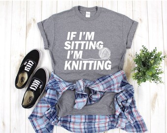 Puede incluir: Camiseta gris con texto blanco que dice "Si estoy sentado estoy tejiendo" y una ilustración blanca de un ovillo de lana y agujas de tejer.