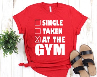 May include: Red t-shirt with white text that reads "Single, Taken, At the Gym" with a checkmark next to "At the Gym".