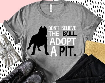 Op de afbeelding: Grijs T-shirt met een zwart silhouet van een pitbull en de tekst "Geloof de bull niet. Adopteer een pit."