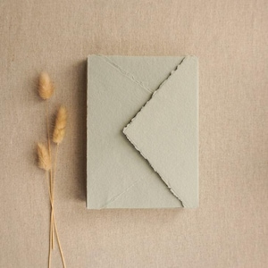 May include: A rectangular envelope made of thick, light sage green paper with a distinct deckled edge on the flap. The envelope is closed and features a textured, fibrous surface. Three dried, tan-coloured grass stalks are positioned to the left.