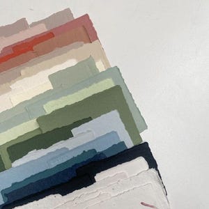 May include: A selection of handmade paper sheets in a range of colours, including pinks, greens, and blues. The paper edges have a rough, torn texture. The papers are stacked diagonally against a white background, creating a layered effect.