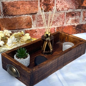 Op de afbeelding: Een rustieke houten dienblad met een donkerbruine afwerking. Het dienblad heeft een kleine pot met een succulent, een zwarte steen, een glazen fles met diffuserstokjes en een kleine witte kaars.