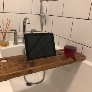 Puede incluir: Una bandeja de baño de madera con un soporte integrado para tableta, un portavasos de vino y dos portavelas. La bandeja es marrón y tiene un acabado rústico. Se coloca en una bañera blanca.