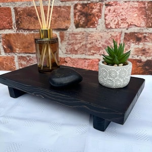 May include: A black wooden tray with a small succulent plant in a white pot, a black stone, and a glass bottle with reeds. The tray is raised on two small legs.