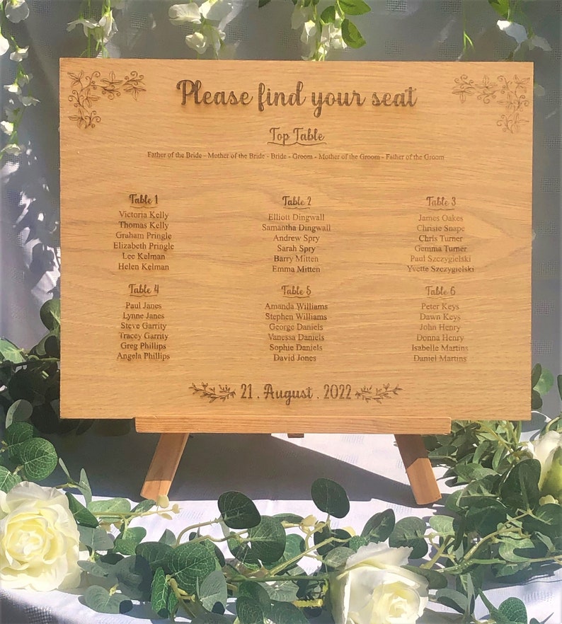 Puede incluir: Un cartel de madera para la asignaci&oacute;n de asientos con el texto "Please find your seat" y "Top Table" en la parte superior. El cartel enumera los nombres de los invitados para cada mesa, con los n&uacute;meros de mesa del 1 al 6. La fecha "21. August 2022" est&aacute; impresa en la parte inferior.