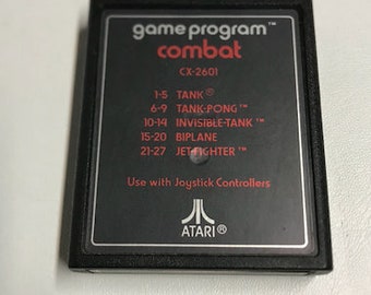 May include: Black Atari game cartridge with the text "game program™ combat CX-2601" and a list of games included: "1-5 TANK®, 6-9 TANK-PONG™, 10-14 INVISIBLE-TANK™, 15-20 BIPLANE, 21-27 JET FIGHTER™". The text "Use with Joystick Controllers" and the Atari logo are also visible.