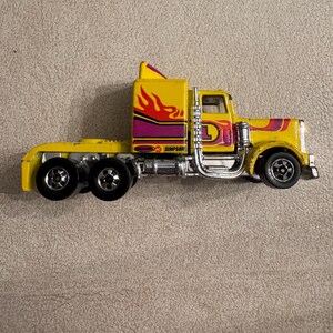 May include: A yellow toy semi-truck with a flame design in red and orange on the cab. The truck has a purple stripe and the text "SIMPSON" is visible. The wheels are black and the exhaust pipe is chrome.