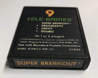 May include: A black Atari game cartridge with a green label that reads "9 TELE-GAMES" and "SUPER BREAKOUT". The cartridge also includes the text "for 1 or 2 players" and "Use with Standard Paddle Controllers".