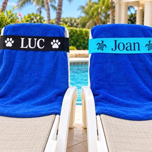 May include: Two bright blue beach towels draped over white lounge chairs. One towel features a black band with the white text LUC and paw prints. The other towel has a light blue band with the dark text Joan and sea turtle graphics.